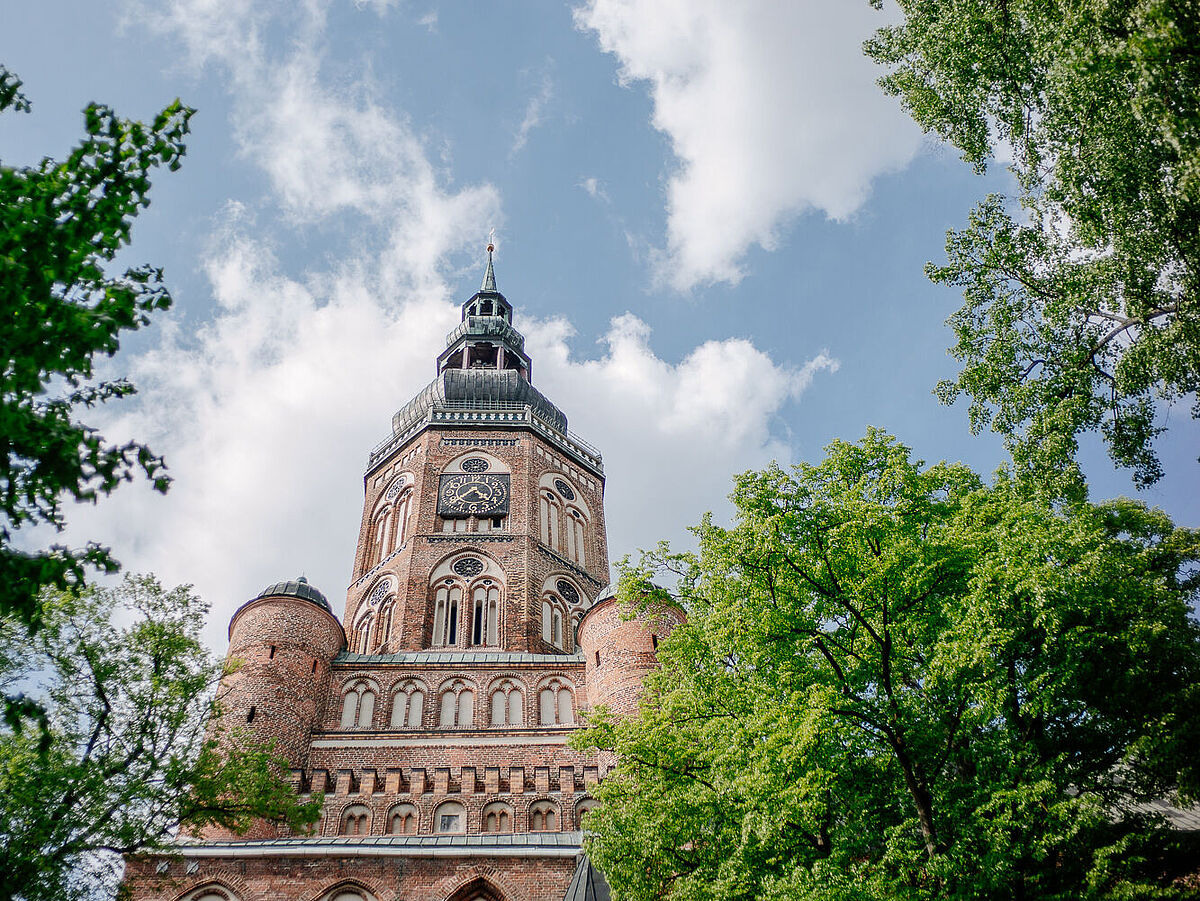 Blick auf den Turm des Domes St. Nikolai. Rechts und links stehen hohe Bäume mit grünem Laub..