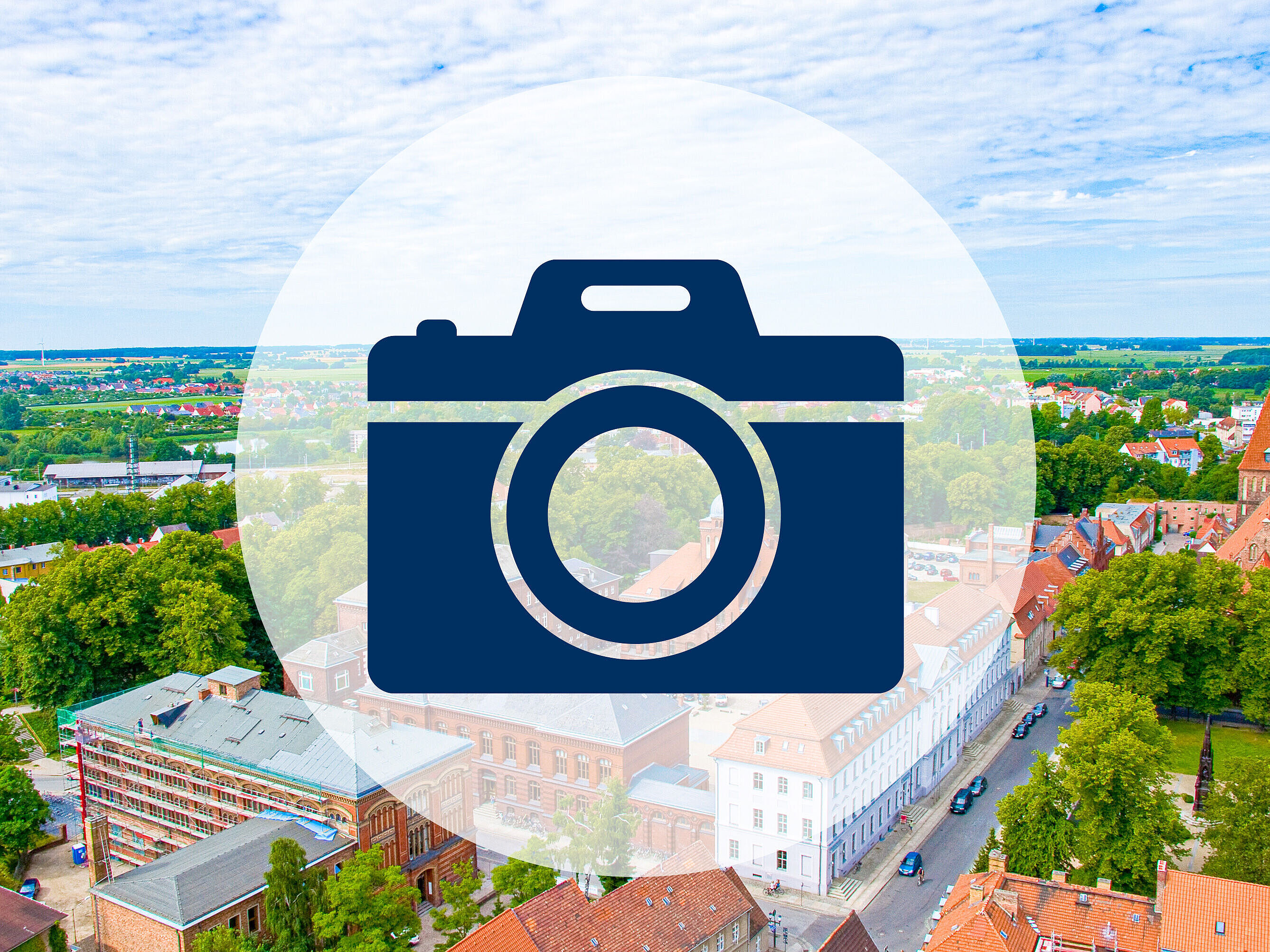 Forschung im Blick Auf einer freundlich hellen Luftaufnahme des historischen Campus der Uni Greifswald ist ein blaues Icon einer Fotokamera.