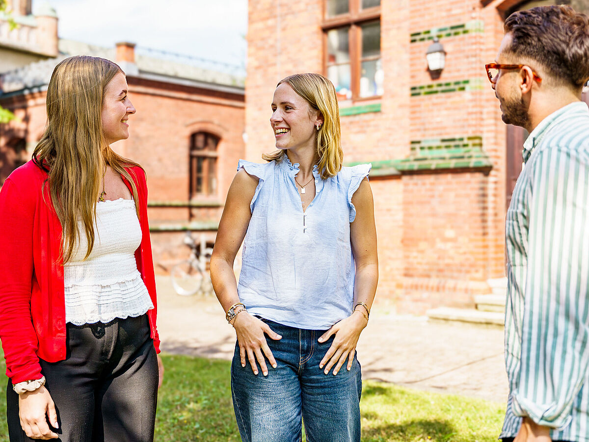 Eine Gruppe von zwei Studentinnen und einem Student stehen bei Sonnenschein vor dem Backsteingebäude der Germanistik und unterhalten sich.