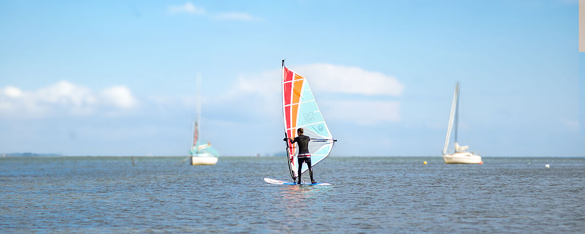 Auf dem Wasser surft ein Mensch mit seinem Segel bei blauem Himmel.