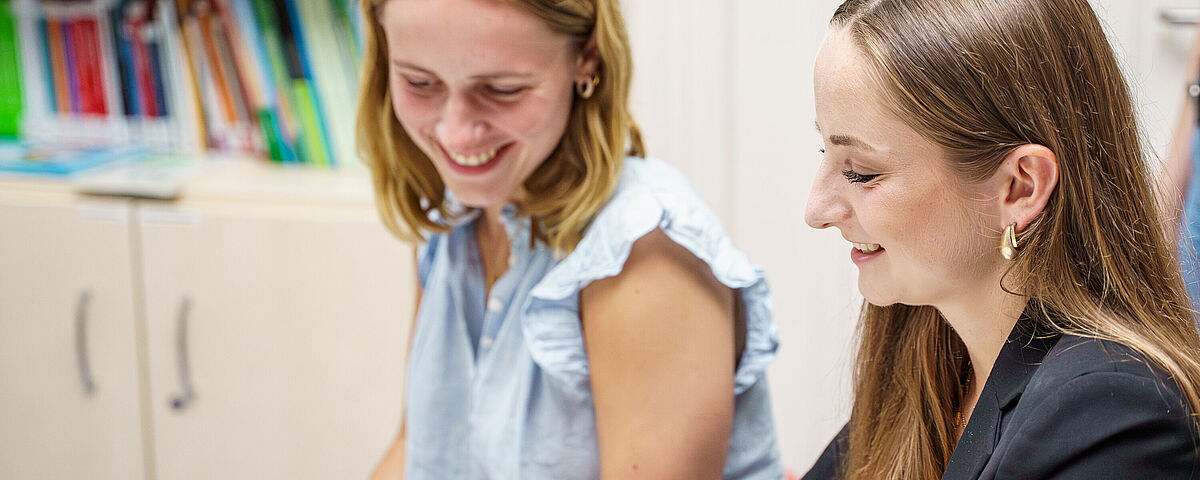 Zwei weibliche Studierende schauen sich gemeinsam ein Buch an und freuen sind dabei.