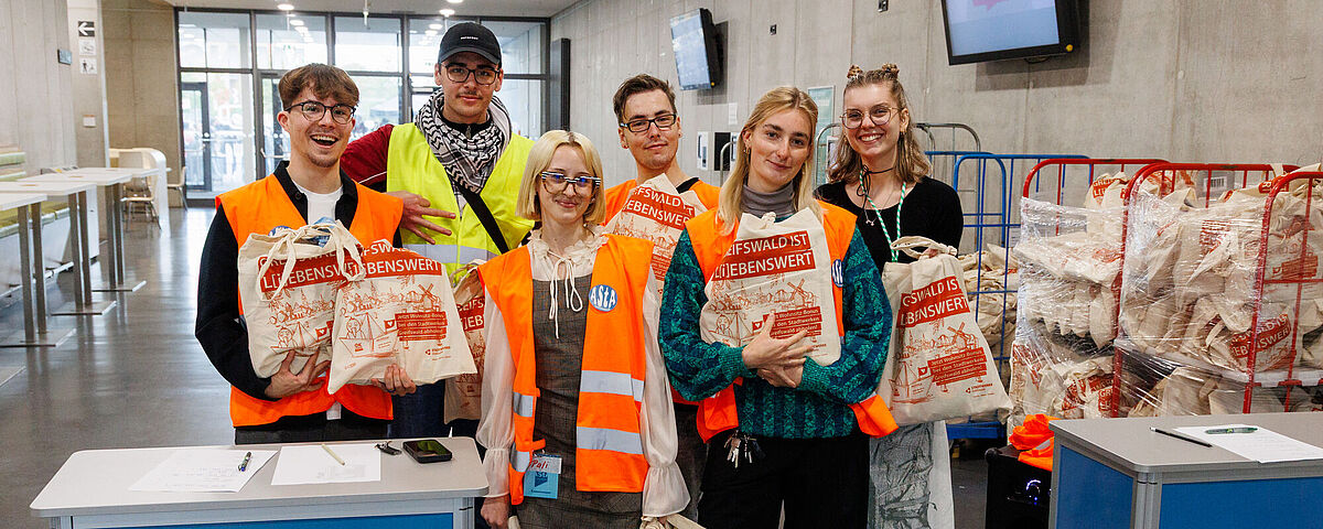 Eine Gruppe von Studierenden schaut erwartungsvoll in die Kamera. Sie haben Ersti-Beutel in der Hand, die sie an die neuen Studierenden verteilen möchten.
