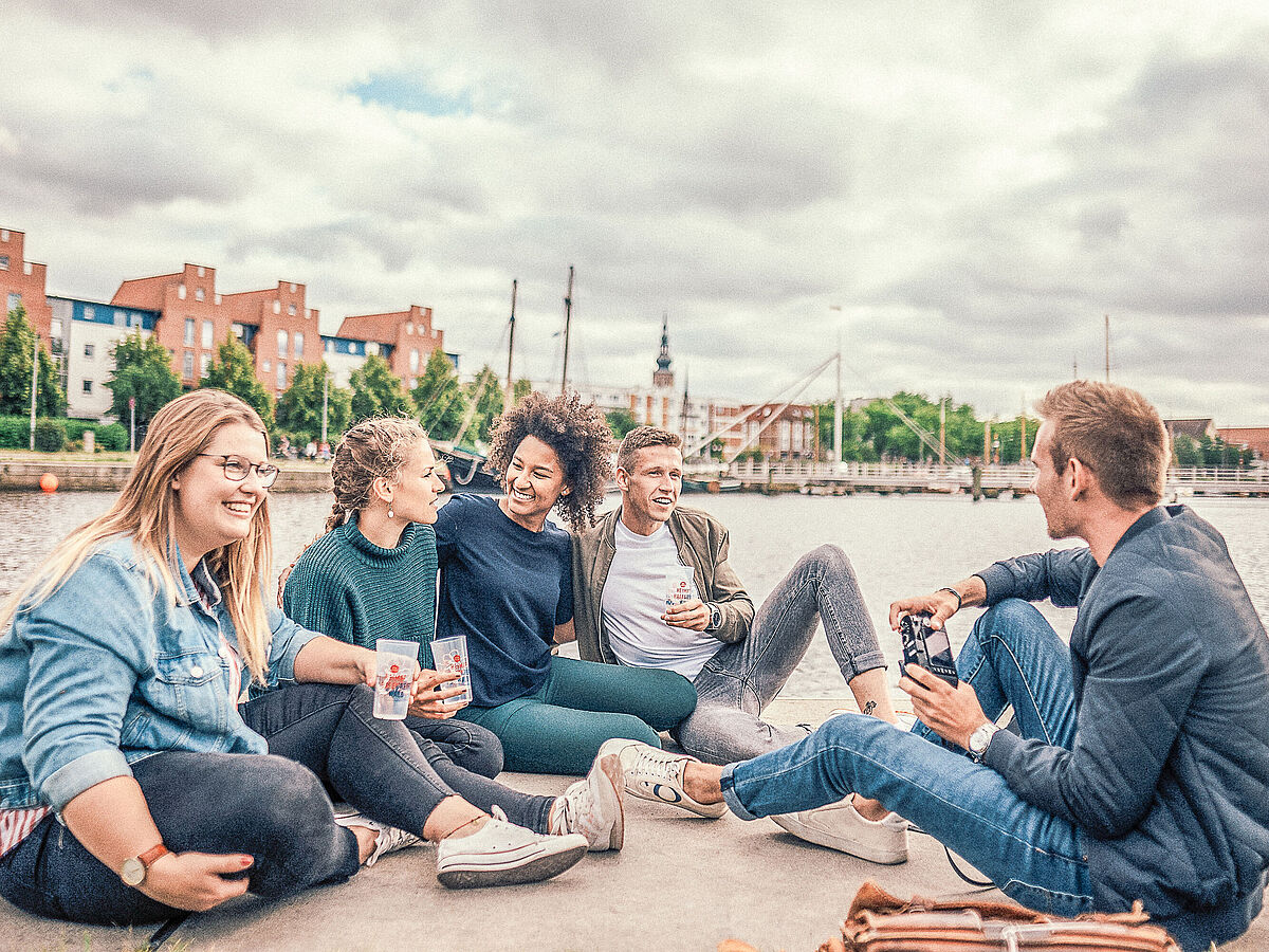 Eine Gruppe Studierender sitzt am Museumshafen am Wasser und unterhält sich angeregt. Im Hintergrund erkennt man neben den historischen Schiffen die Stadt Greifswald mit dem Dom. 
