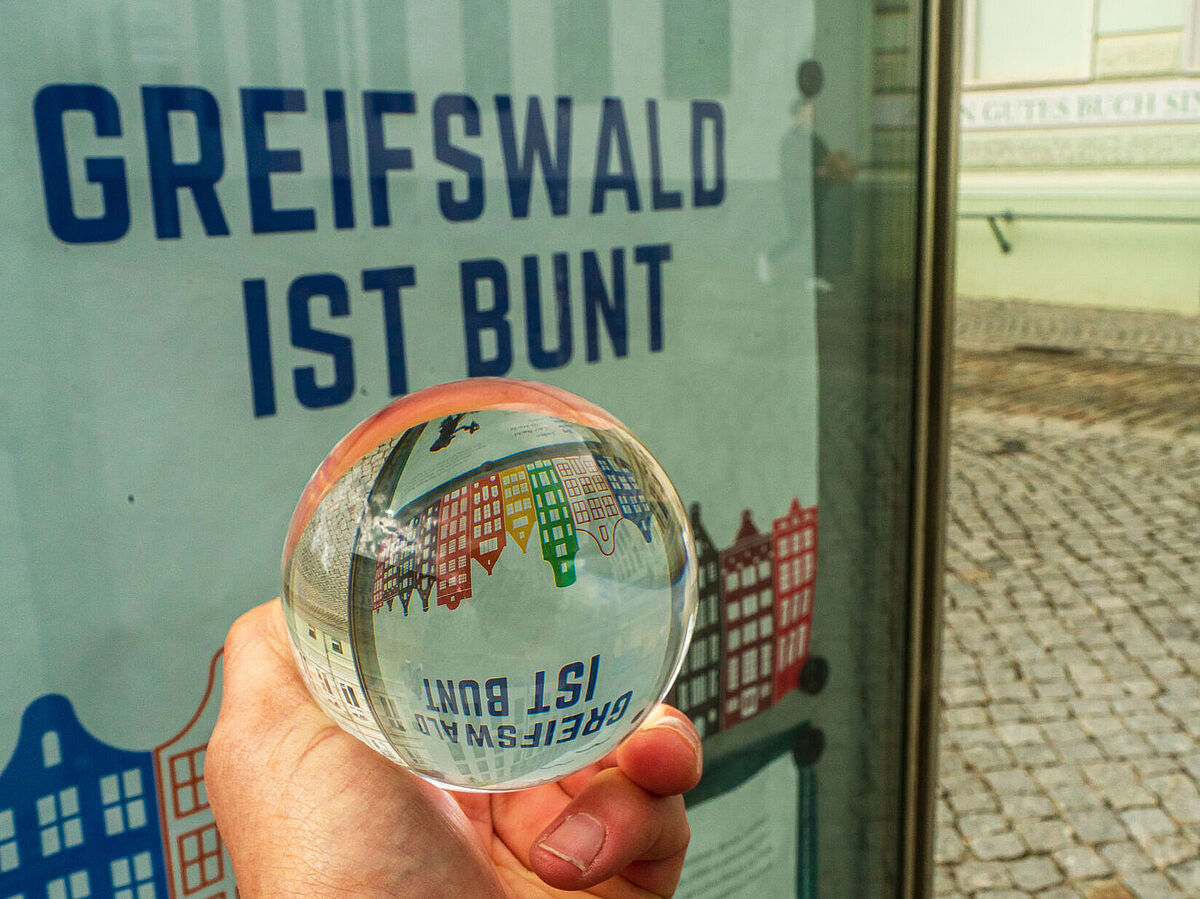 A hand holding a crystal ball in front of a poster with the words "Greifswald ist bunt"